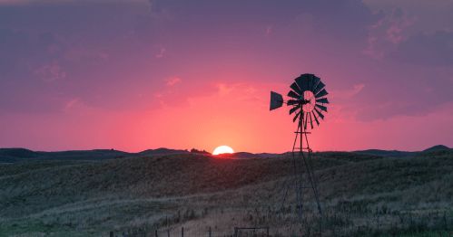 A windmill silhouette stands on a grassy hill against a vibrant pink and orange sunset.