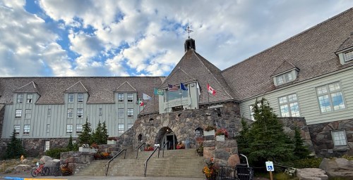A historic lodge with a massive stone façade soars into a blue sky with white clouds.