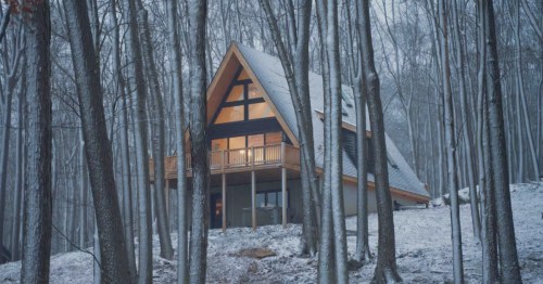 A-frame cabin with a raised deck, nestled in a snowy forest with tall, bare trees.