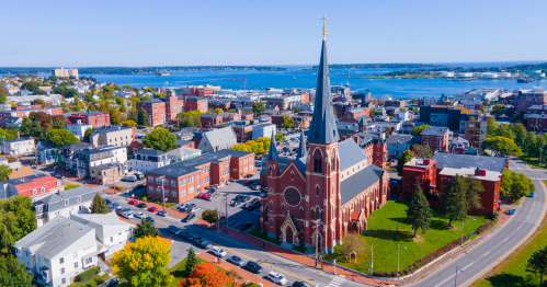 Aerial view of downtown Portland, showcasing historic brick church and blue bay under a light blue sky in the summertime.