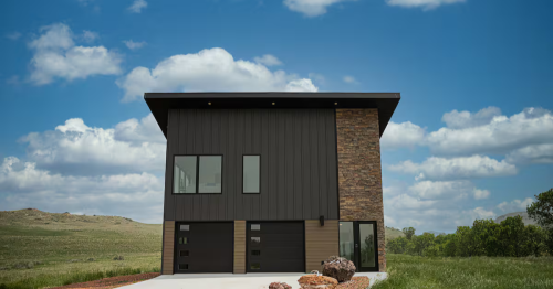 Modern, two-story vacation rental with dark siding and a stone accent against a backdrop of blue sky and green hills.