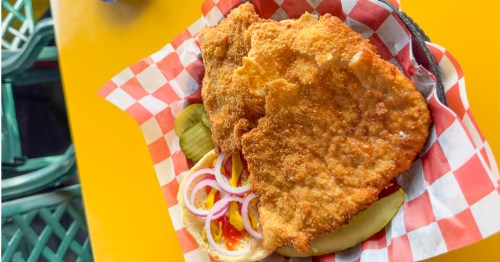 A pork tenderloin sandwich with onions and pickles in a basket with red and white checkered paper on top of a yellow table