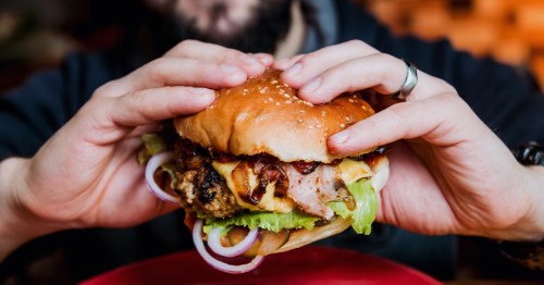 A person holds a large cheeseburger with bacon, lettuce, and tomato between his two hands.