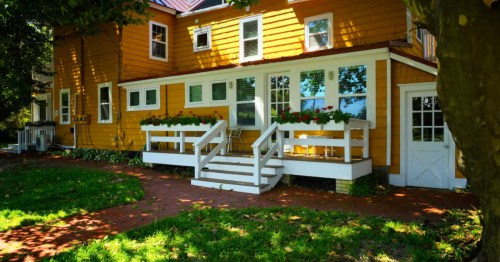 The exterior of a charming yellow cottage surrounded by a canopy of trees.