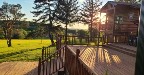A large wooden cabin facade at sunset, with a porch overlooking a green park.