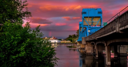 A bright blue bridge sits across a calm river with a fiery sunset overhead.