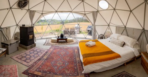 Wide interior view of the Sunset Dome showing the bed, rugs, wood stove, and a panoramic view of the fields outside.