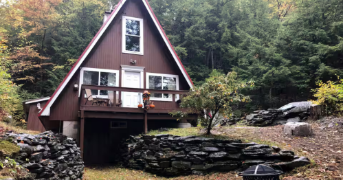 The exterior of the Berkshire A-Frame Cabin AIrbnb property in Cummington, Massachusetts. The brown cabin is surrounded by large green trees.