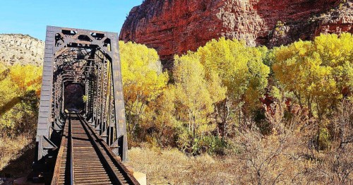 Iron train bridge leading into a red rock canyon in Arizona, framed by vivid yellow cottonwoods and clear blue sky.