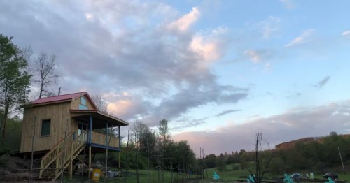 A wide view of a two-story farm cabin next to a vegetable garden, under a clear sky at sunset.