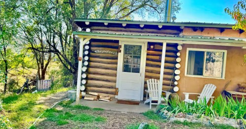 Rustic log cabin in New Mexico surrounded by trees, featuring a small covered porch with two white Adirondack chairs, stacked firewood by the door, and a hand-painted sign reading “D.H. Lawrence’s.”