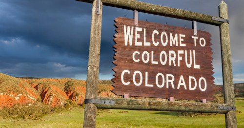 "Welcome to Colorful Colorado" wooden sign in front of green and golden Colorado landscape.