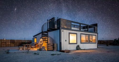 A modern, container-style home with warm interior lights and a spiral staircase leading to a rooftop deck sits under a star-filled night sky in a remote desert setting.