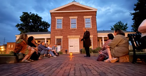 A crowd gathered on a brick street in front of an old, brick building. It's almost dark, a man dressed in black speaks to the crowd with his glowing lantern on the ground near his feet.