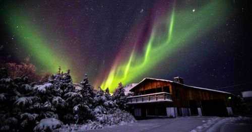 A two-story cabin with a large upper deck, surrounded by snow-flocked trees, with green and purple aurora lights shimmering above it.