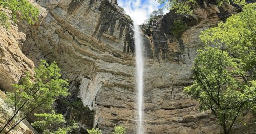 A tall, narrow waterfall streaming gracefully over limestone cliffs surrounded by bright green trees in northern Arkansas.