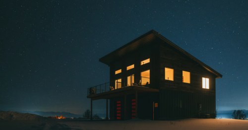 A modern-style Airbnb with a large second-floor deck at night. Vibrant stars shine overhead, and light flows out of the windows.