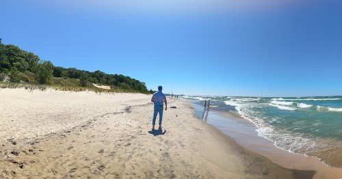 A man standing on a wide, sandy beach between a forested sand dune and turquoise water with waves