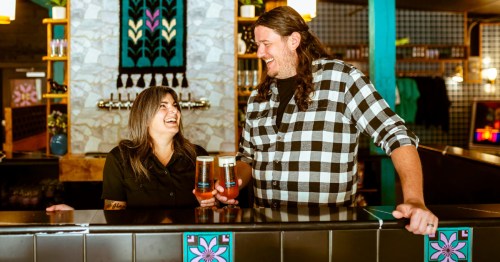 Two people behind counter holding beers