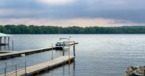 A pontoon boat parked by a woddend dock on a sparkly, inky body of water with tree lined banks on the other side