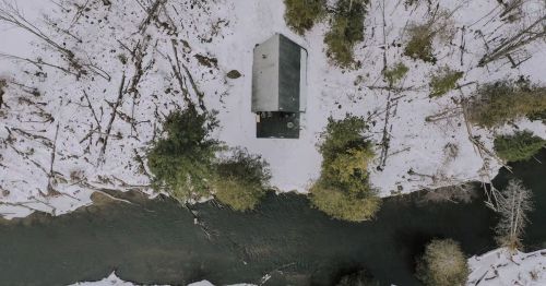 A birds-eye view of an A-frame cabin in a forest along the shores of a river.