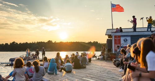 A group of people sitting on a dock at dusk, there's a beautiful golden glow over the sky and water, illumating an American flag
