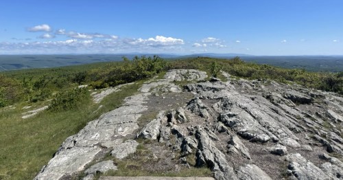 Panoramic view from a rocky ridge, showing distant green mountains under a bright blue sky.