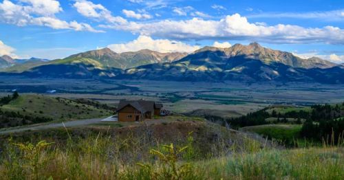 A wide open mountain valley with a cabin perched on a hillside overlooking the rolling foothills and layered blue mountain peaks under a sunny blue sky.