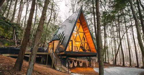 An elevated A-frame with large windows and hammocks underneath in a forest.