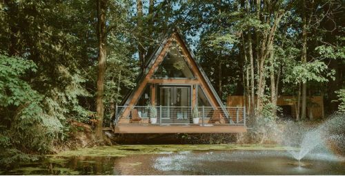 An A-frame home with a patio sitting over a pond with a water fountain.