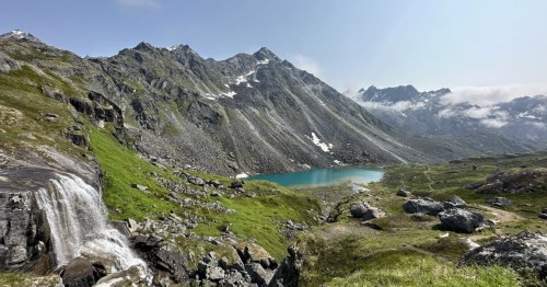 A waterfall tumbles off a small cliff with a large mountain peak and small lake in the distance.