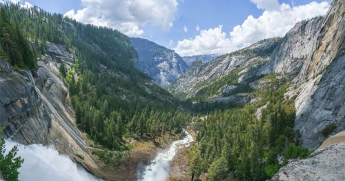 A powerful waterfall cascades down a steep granite cliff into a forested valley surrounded by rugged mountains under a partly cloudy sky.