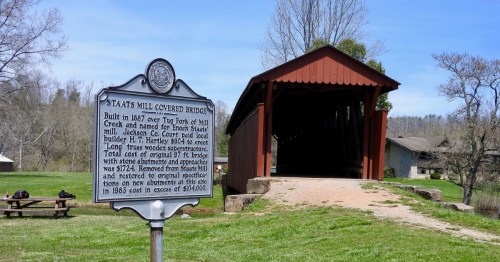 A historic sign next to a red covered bridge on a sunny day.