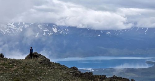 A woman stands on the top of a rocky cliff overlooking water, mountains, and fog on an Alaskan hiking trail.