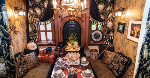 In the middle of an elegant room, there's a long table covered in tea cups and finger foods.