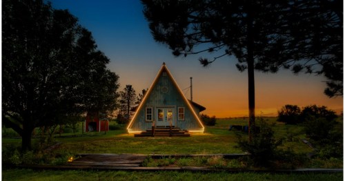 A light blue A-frame cabin, outlined with glowing rope lights, set in a rural field at sunset.
