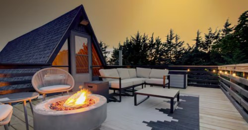 A modern A-frame cabin deck at sunset with a fire pit, sectional sofa, and string lights, surrounded by trees.