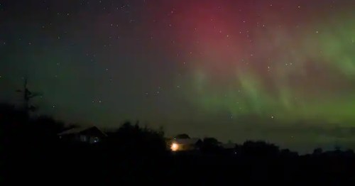 A spectacular view of the Northern Lights with bright green and red bands over a dark, rural horizon. 
