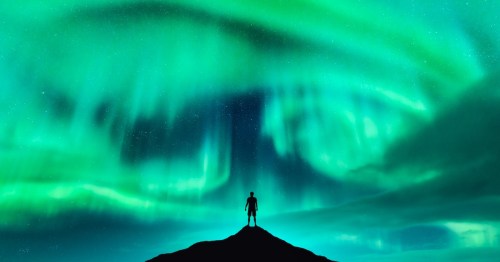 A man stands in silhouette on a mountain peak beneath a swirling maelstrom of green and blue northern lights.