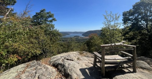 A rustic wooden chair sits on a thick stone slab overlooking a view of a gree forest and wide river.