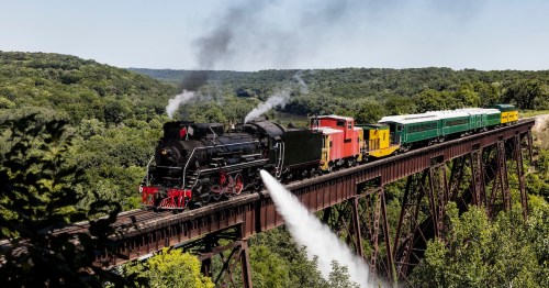 A steam locomotive pulling passenger cars crosses a high trestle bridge over a forested valley.