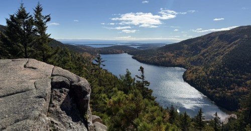 A long lake glistens in the sun between two mountains filled with a forest starting to change for autumn. A sandy cliffside sits int the forefront.