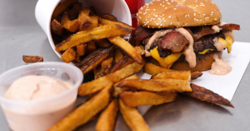 Close-up of a bacon cheeseburger and tipped container of fries on a grey surface