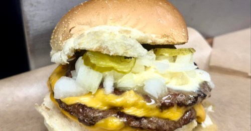 A close-up of a double cheeseburger on a buttered bun, topped with a generous pile of diced raw onions and sliced dill pickles.