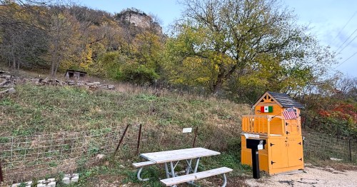 A colorful wooden structure labeled "The Bread Hut" near a road, with an towering limestone bluff in the background.