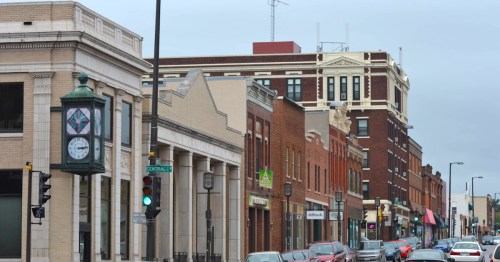 Cars line the bustling street of a historic downtown area with red brick and stone buildings in a small Wisconsin city.