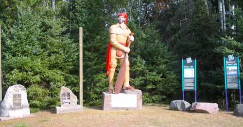 A large statue of a man in historical attire, holding a paddle, surrounded by trees and a stone base with inscriptions.