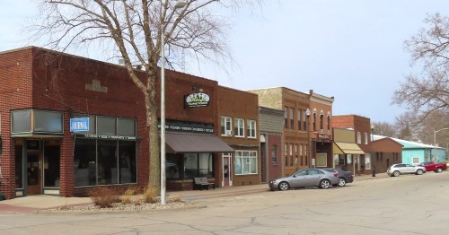 A quaint street scene featuring shops, a newspaper office, and parked cars in a small town.
