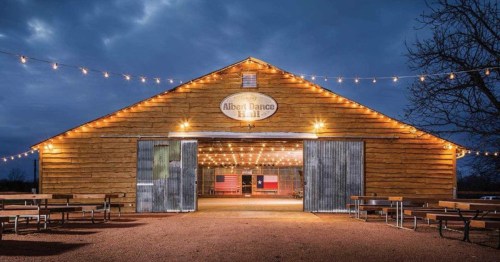 exterior of the Albert dancehall with open doors and picnic tables outside along with string lights at night in albert Texas