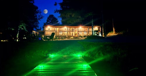 Exterior of the Mystic Pond Cabin lit up at night and in the foreground is a footbridge with glowing green lights and a full moon in the sky